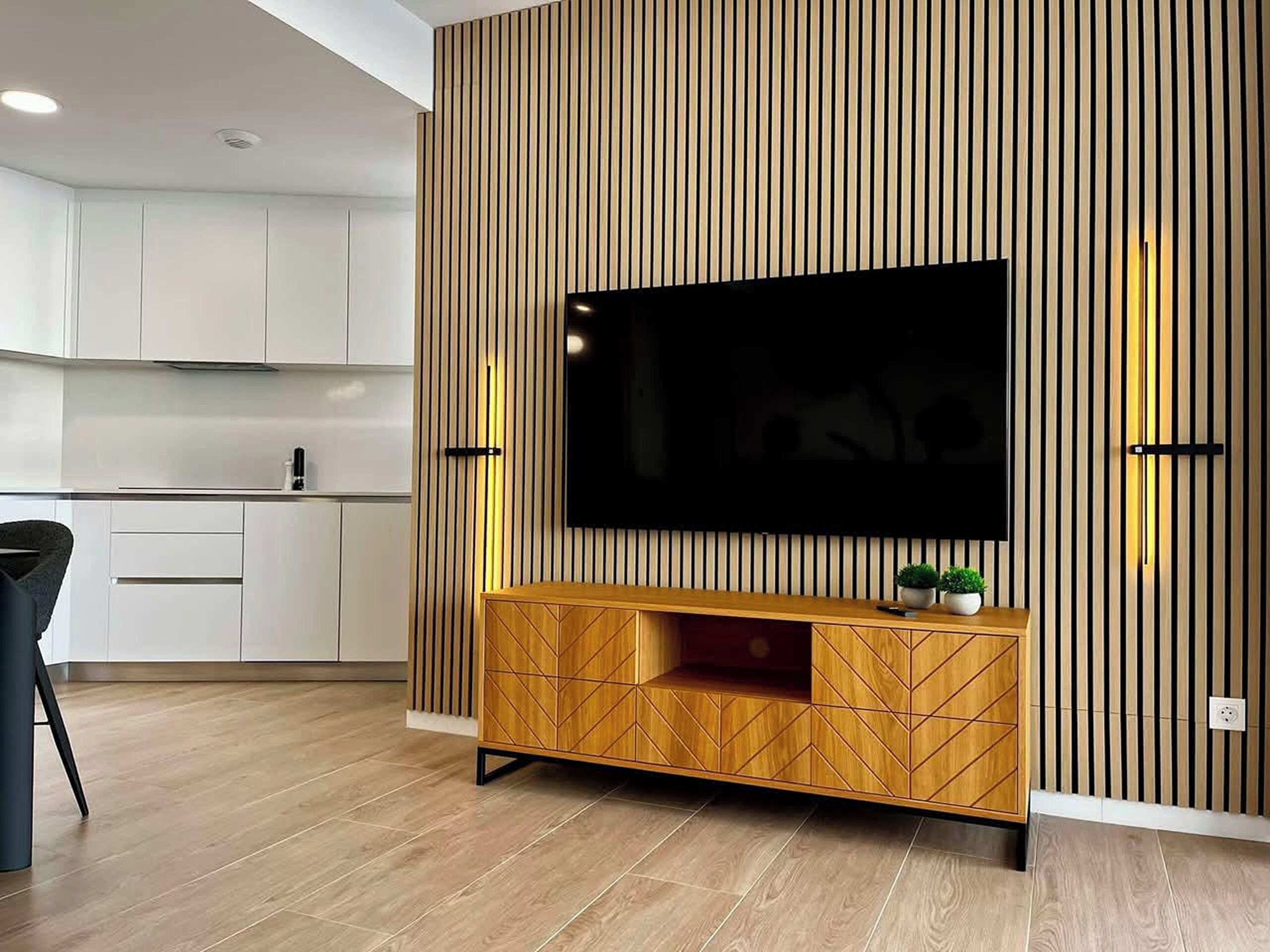 A modern living room with a flat-screen TV mounted on a wooden wall, a wooden cabinet below, and a kitchen area with white cabinets in the background.