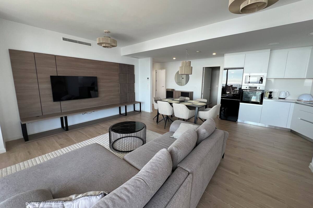 Una sala de estar moderna con un televisor de pantalla plana grande, un sofá gris, una zona de comedor con mesa y sillas, y una cocina con armarios y electrodomésticos blancos.