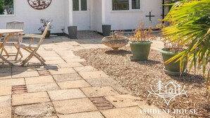 Een wit gebouw met een rond logo op de muur, een terras met een tafel en stoelen, en planten in potten.