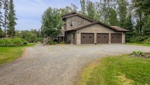A two-story wooden building with a garage, surrounded by a gravel driveway and lush greenery. - Kenai Riverside Escape (Soldotna)