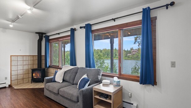 A room with a sofa, a small table, a heater, and large windows with curtains. - Kenai Riverside Escape (Soldotna)