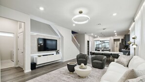 Amplia sala de estar con una televisión de pantalla plana grande, un sofá blanco y un comedor, con una cocina al fondo.