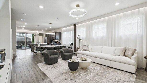 Un amplio salón con un sofá blanco, dos sillones y una mesa de centro. Se ve una cocina al fondo con electrodomésticos modernos.