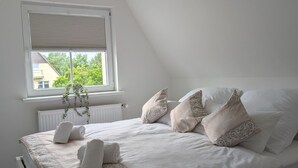 Ein Schlafzimmer mit einem weißen Bett, einem gemusterten Bettüberwurf und dekorativen Kissen. Es gibt ein Fenster mit Jalousien, eine Heizung und eine Pflanze auf dem Fensterbrett.
