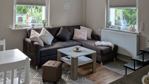 Ein gemütliches Wohnzimmer mit einer Eckcouch, einem kleinen Couchtisch und einem Esstisch. Zwei Fenster mit Jalousien, ein Heizkörper und eine Deckenleuchte sind vorhanden.