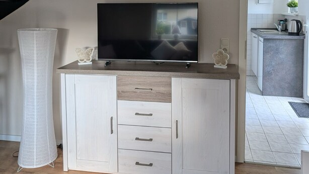 Ein modernes Wohnzimmer mit Flachbildfernseher auf einem Holzschrank, einer Treppe und Blick in die Küche.