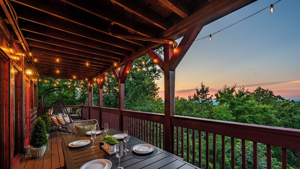 Una terraza de madera con una mesa lista para comer, rodeada por una barandilla y luces colgantes, con vistas a un área boscosa.