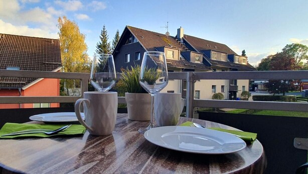 Una terraza con una mesa dispuesta para dos, que incluye una copa de vino, una taza y un plato, con vistas a casas al fondo.