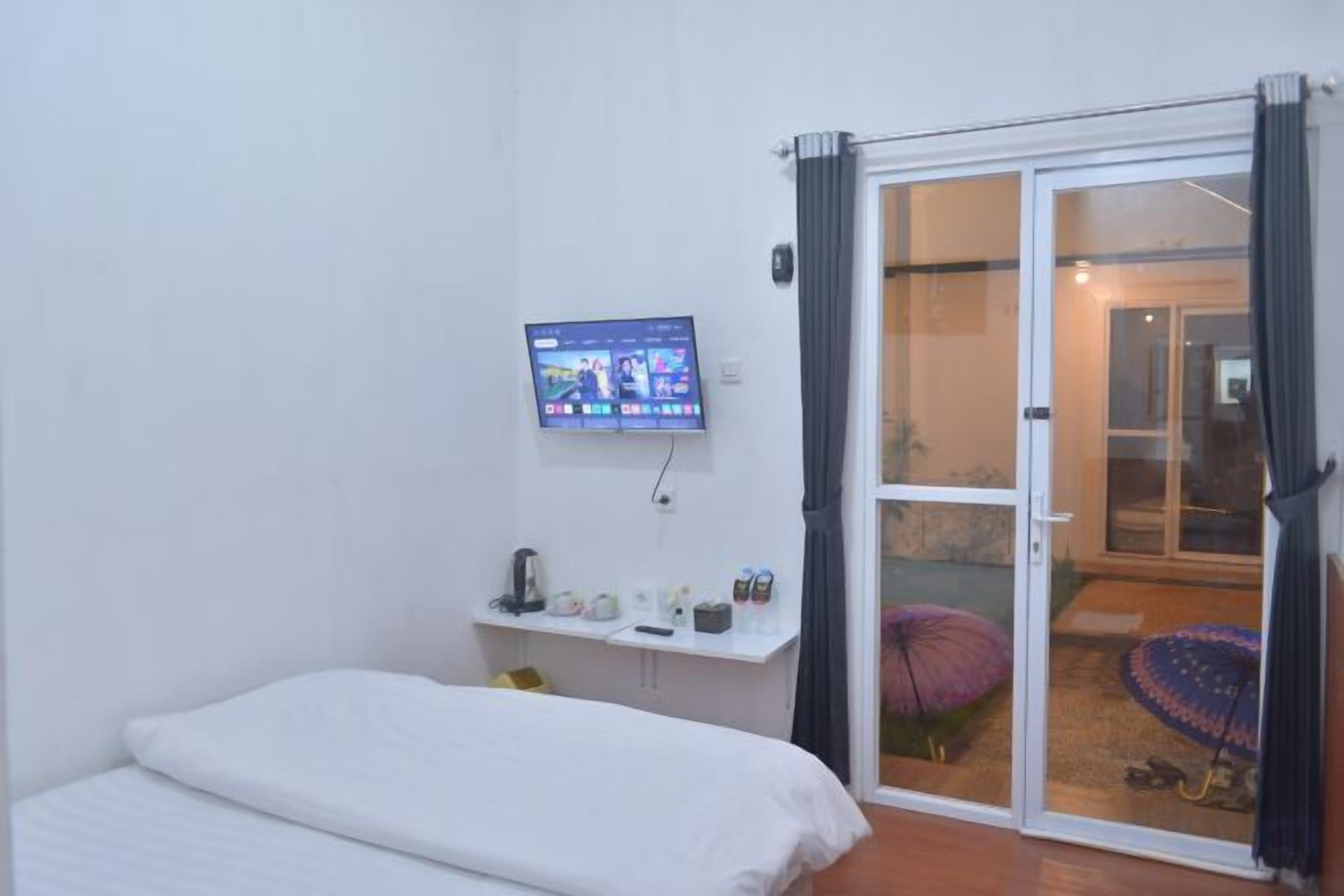 A bedroom with a bed, a TV mounted on the wall, a small shelf with items, and a sliding glass door leading to a balcony.