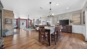 Un vaste salon doté d’un plancher en bois franc, un coin repas avec une table et des chaises, un grand téléviseur à écran plat et une cheminée.