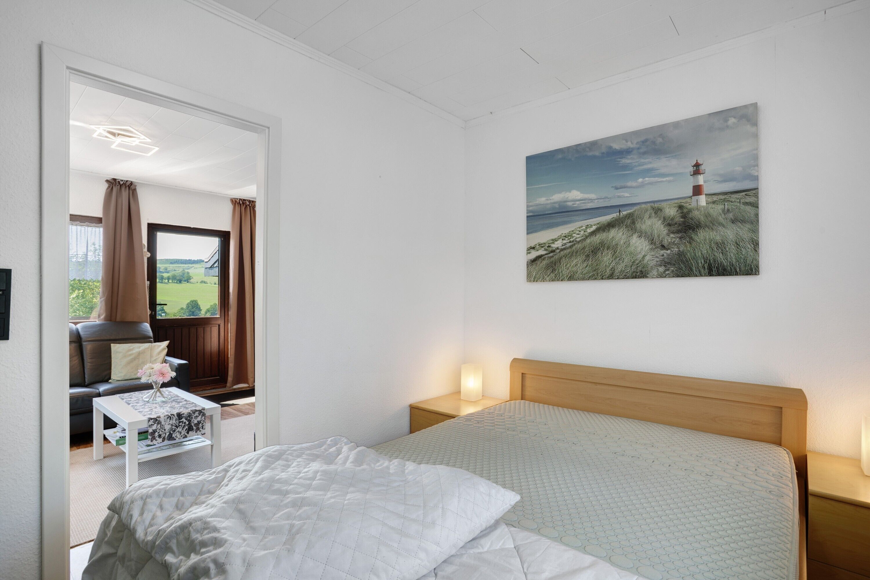 Ein Schlafzimmer mit Bett, Nachttisch mit Lampe, einem Leuchtturm-Gemälde und Blick auf einen Garten durch eine offene Tür.