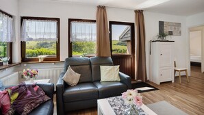 A living room with a leather sofa, a white cabinet, and a view of greenery through large windows. - Apartment 'Bianca' with Lake View, Private Terrace and Wi-Fi (Meschede)