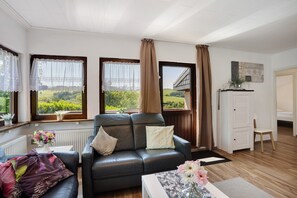 A living room with a leather sofa, a white cabinet, and a view of greenery through large windows. - Apartment 'Bianca' with Lake View, Private Terrace and Wi-Fi (Meschede)
