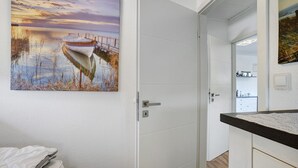A room with a white door, a painting of a boat on water, a bed with a white cover, and a small desk with a lamp. - Apartment 'Bianca' with Lake View, Private Terrace and Wi-Fi (Meschede)