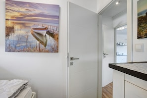 A room with a white door, a painting of a boat on water, a bed with a white cover, and a small desk with a lamp. - Apartment 'Bianca' with Lake View, Private Terrace and Wi-Fi (Meschede)