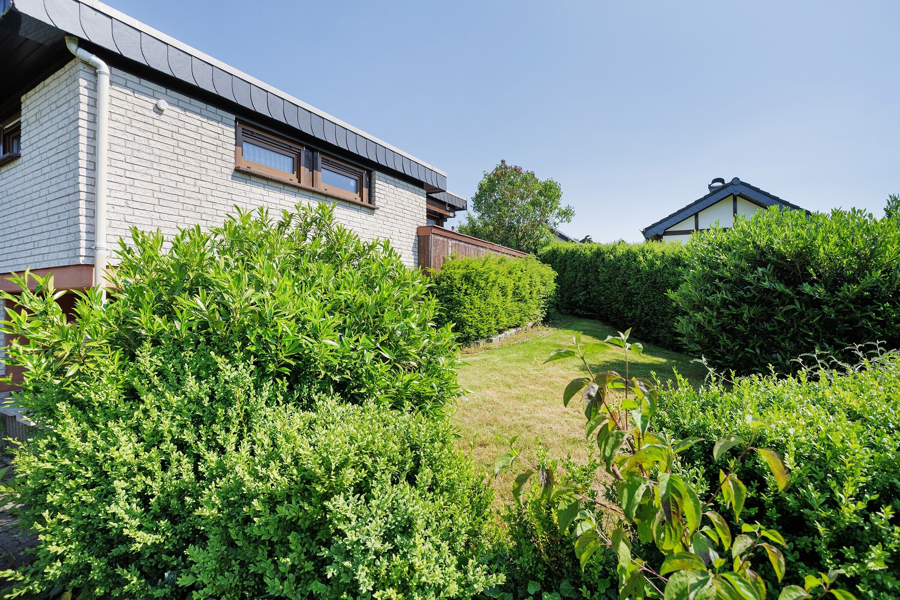 Ein Wohngebäude mit Backsteinfassade, einem geneigten Dach und einem gepflegten Garten mit verschiedenen Sträuchern und Rasen.