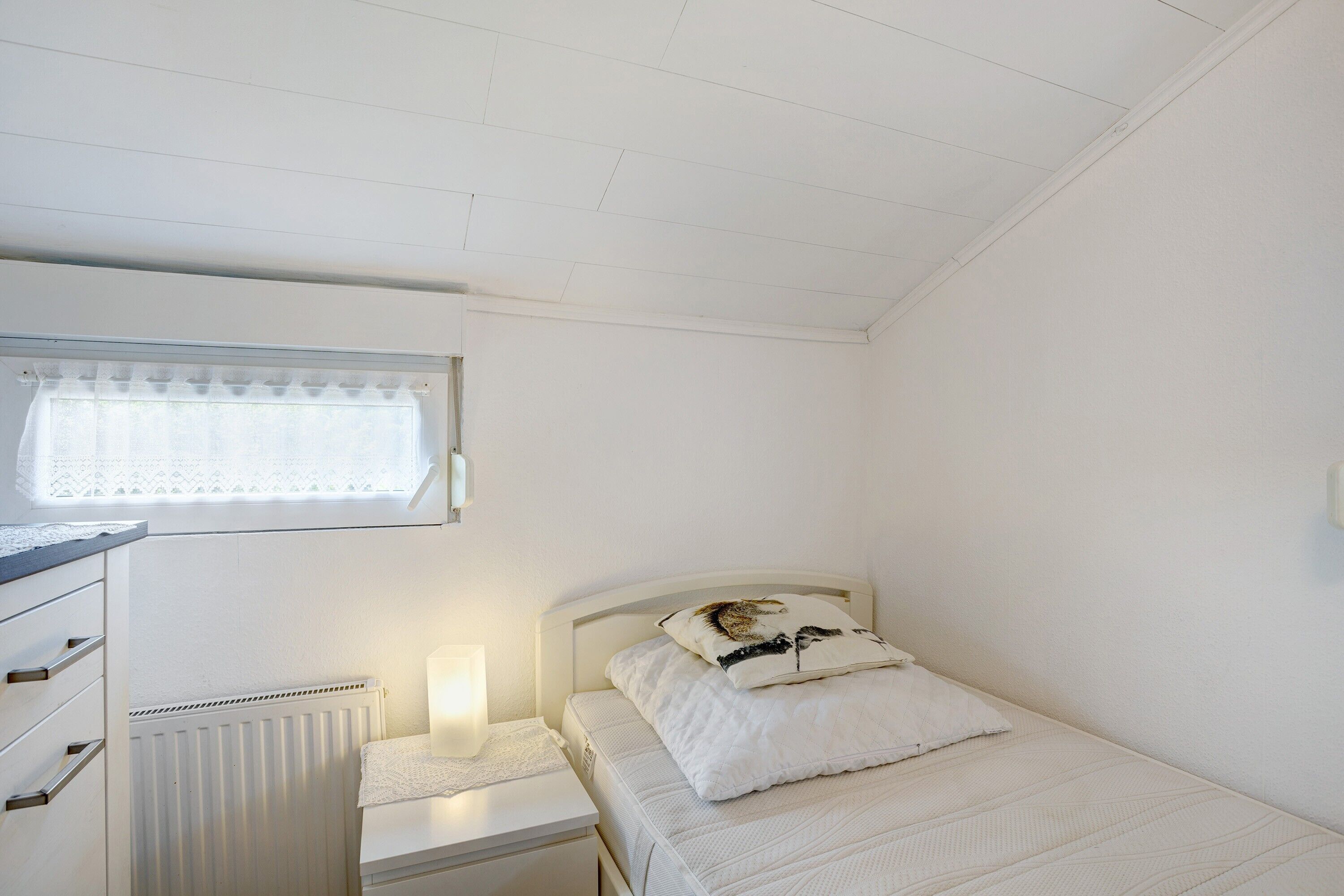 A bedroom with a single bed, a nightstand, a lamp, a radiator, and a window with a curtain.