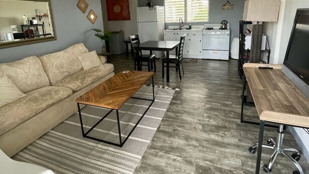 A spacious living room with a sofa, coffee table, and TV stand. A kitchen area with a dining table and chairs is visible in the background. - Charming apartment with AC in Independence/Mango Creek. Near Placencia hot spot! (Independence and Mango Creek)
