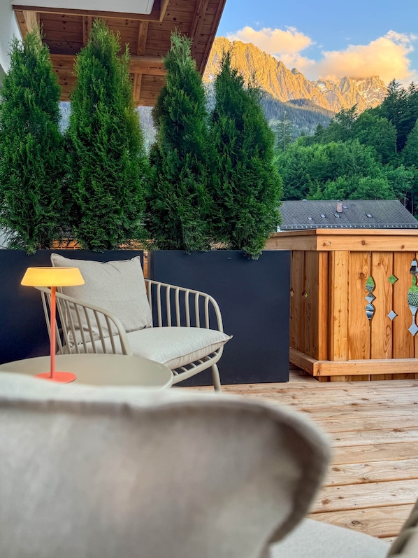 Eine Holzterrasse mit einem weißen Metallstuhl und einer Tischlampe, mit Blick auf eine Berglandschaft.