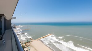 A view from a balcony overlooking a beach with waves, a clear sky, and a distant city skyline.