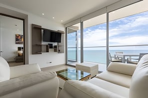 A modern living room with a large window overlooking the sea, a flat-screen TV mounted on the wall, and a glass coffee table.