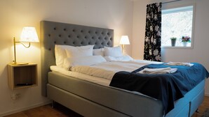 Una cama bien hecha con sábanas blancas y una manta azul oscuro, dos lámparas de noche, un cabecero tapizado en gris y una ventana con cortinas florales.