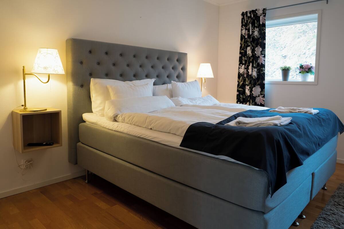 Una cama bien hecha con sábanas blancas y una manta azul oscuro, dos lámparas de noche, un cabecero tapizado en gris y una ventana con cortinas florales.