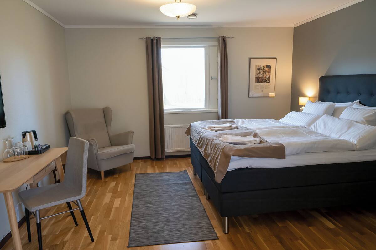 Habitación de hotel con cama, escritorio con silla, cafetera y ventana con cortinas.
