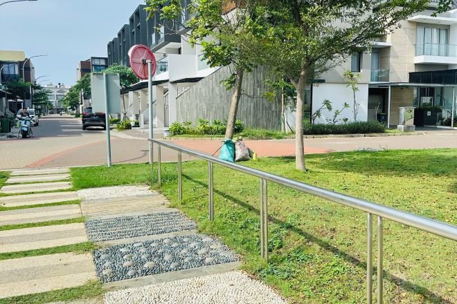 A residential street with a sidewalk, a metal railing, and a grassy area with trees.