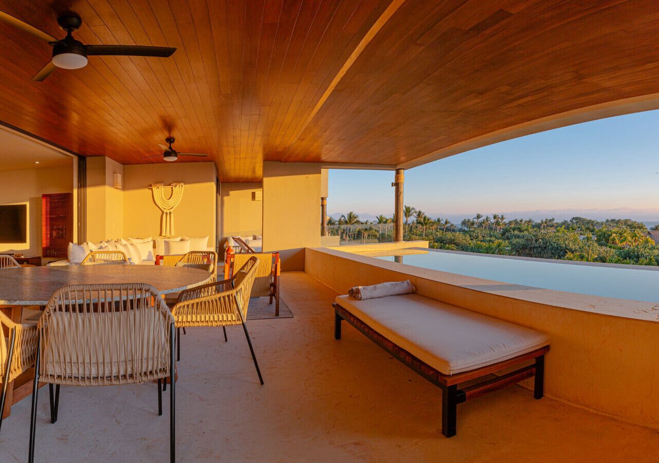 A spacious outdoor area with a wooden ceiling, a dining table with chairs, a sofa, and a view of a lush landscape.