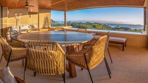 A round dining table with wicker chairs, a bench, and a view of a coastal landscape. - Condo Haramara Luxury Oceanview in TAU Punta Mita (Punta Mita)