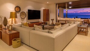 A modern living room with a large sectional sofa, a flat-screen TV, and a view of the ocean at dusk. - Condo Haramara Luxury Oceanview in TAU Punta Mita (Punta Mita)