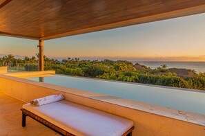 Een moderne lounge buiten met een zwembad, een houten bank en uitzicht op de oceaan en een tropisch landschap.