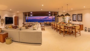 A modern living room with a large sofa, dining area with wooden chairs, and a view of the ocean through a large window. - Condo Haramara Luxury Oceanview in TAU Punta Mita (Punta Mita)