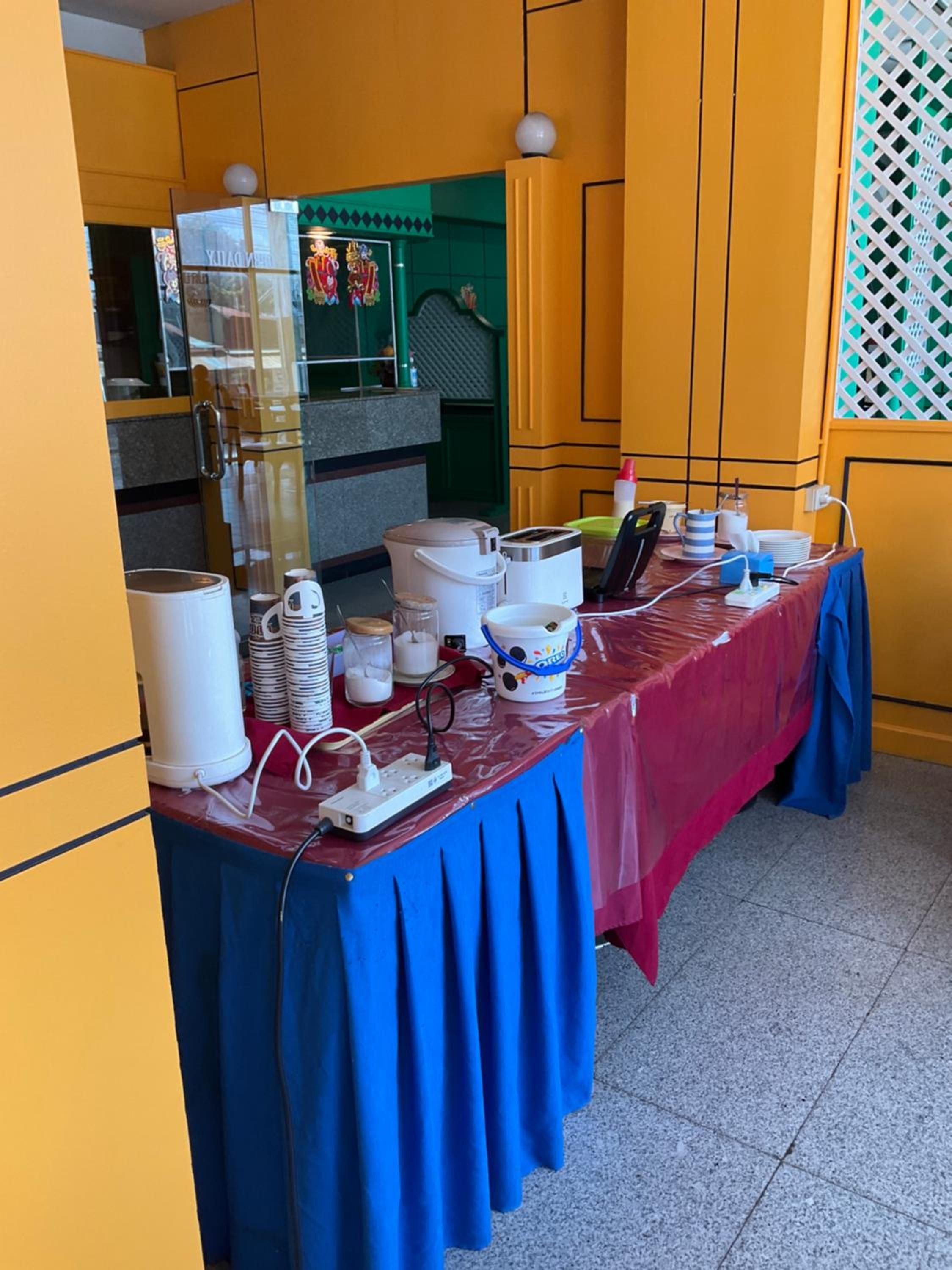A table set up with various kitchen appliances and supplies, a power strip, and a blue tablecloth in a room with yellow walls and a green door.