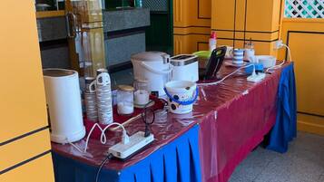A table set up with various kitchen appliances and supplies, a power strip, and a blue tablecloth in a room with yellow walls and a green door.