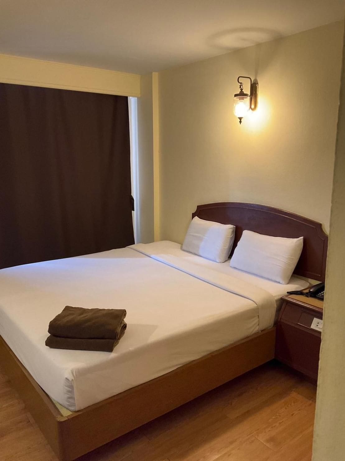 Een enkel bed met houten frame, witte beddengoed en een bruine deken die netjes op het bed is opgevouwen. Er staat een nachtkastje met een telefoon en een lamp.