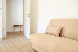 Un canapé beige avec un coussin, une chaise en bois et une porte blanche dans une pièce dont le sol est recouvert de parquet clair.