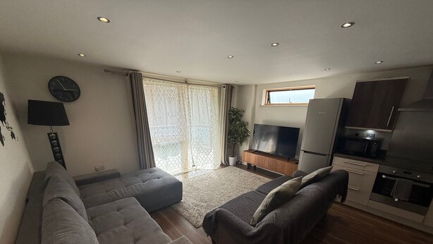 A modern living room with a sectional sofa, a flat-screen TV, and a kitchen area with a refrigerator and microwave.