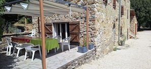 A rustic outdoor seating area with stone walls, a table with a green tablecloth, and white plastic chairs.