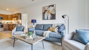 Una sala de estar moderna con sofá, sillones y una mesa de centro. Se observa una cocina al fondo con armarios de madera y un microondas.