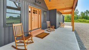 Een overdekt terras met houten schommelstoelen en een bank, een houten deur en een raam met uitzicht op een groen landschap.