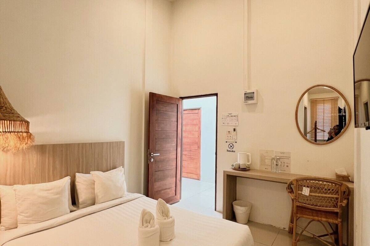 Habitación de hotel con una cama grande, una mesita de noche con espejo, una silla de madera y un escritorio con computadora y una pequeña estantería.