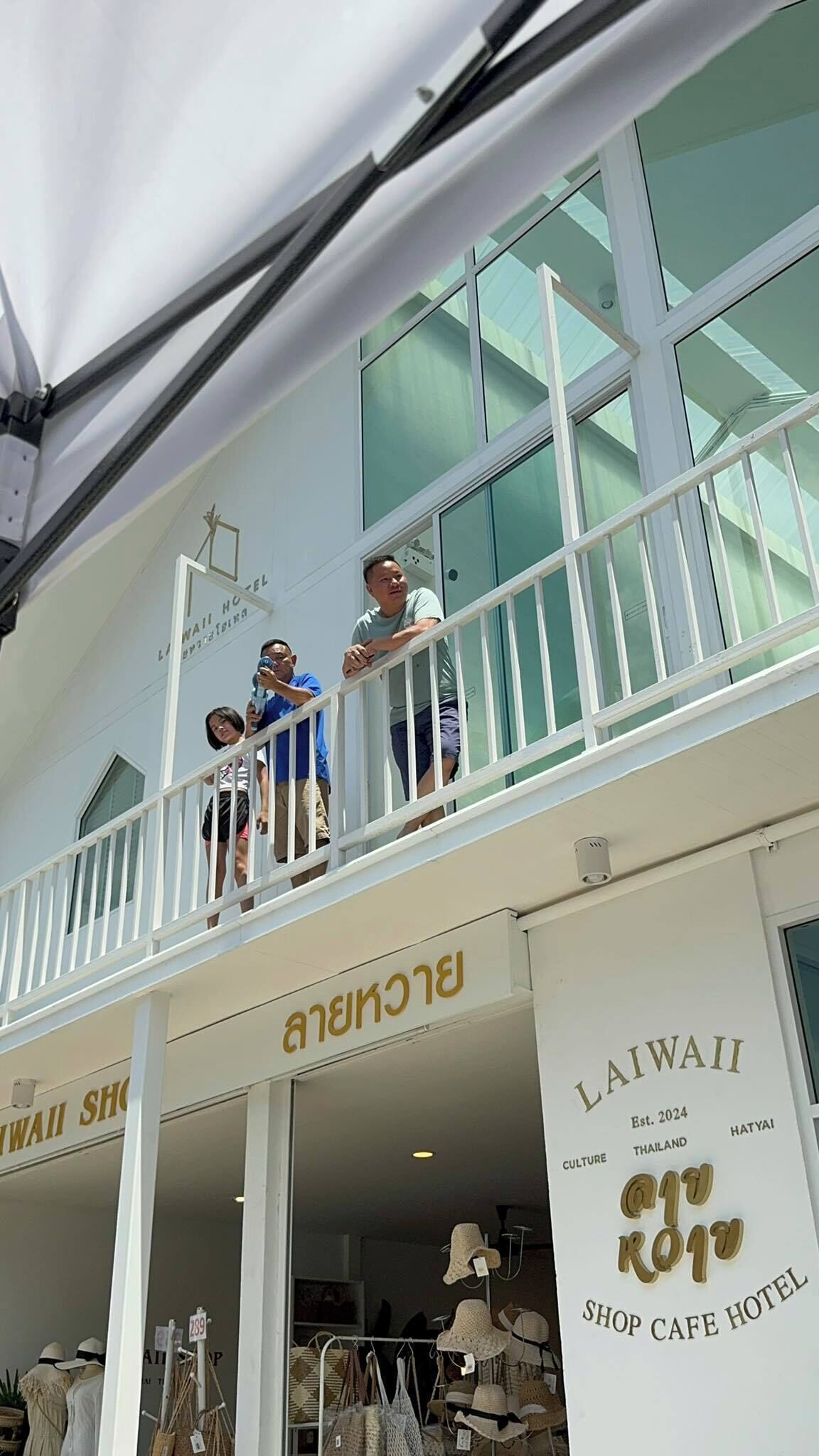 Un bâtiment à deux étages doté d’un balcon. Au rez-de-chaussée, on peut voir une enseigne indiquant « LAWAII SHOP CAFE HOTEL », avec la mention « Fondé en 2024 » et le lieu « THAÏLANDE, HATAYI ». L’étage supérieur porte une enseigne indiquant « BLACKBIRD ».