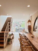 A modern interior with a wooden staircase, a dining area with wicker chairs, and a large window offering a view of the street.
