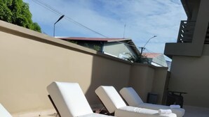 Une rangée de chaises longues dotées de coussins blancs et de serviettes, disposées sur une terrasse carrelée, avec un mur de couleur beige et un bâtiment à toit rouge en arrière-plan.