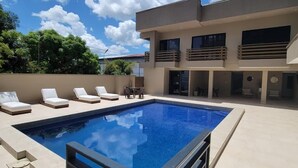 Un espace piscine moderne doté d’une piscine rectangulaire, de chaises longues et d’un bâtiment à deux étages avec des balcons.