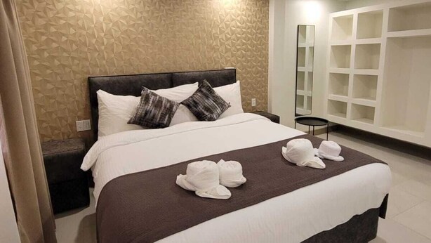 Un lit bien fait, avec du linge de lit blanc et marron, une tête de lit sombre et deux oreillers. On peut voir une couverture marron et deux serviettes blanches sur le lit.
