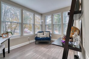 A room with a blue sofa, a small table, and a lamp. There are large windows with blinds, and a plant is visible on the windowsill.