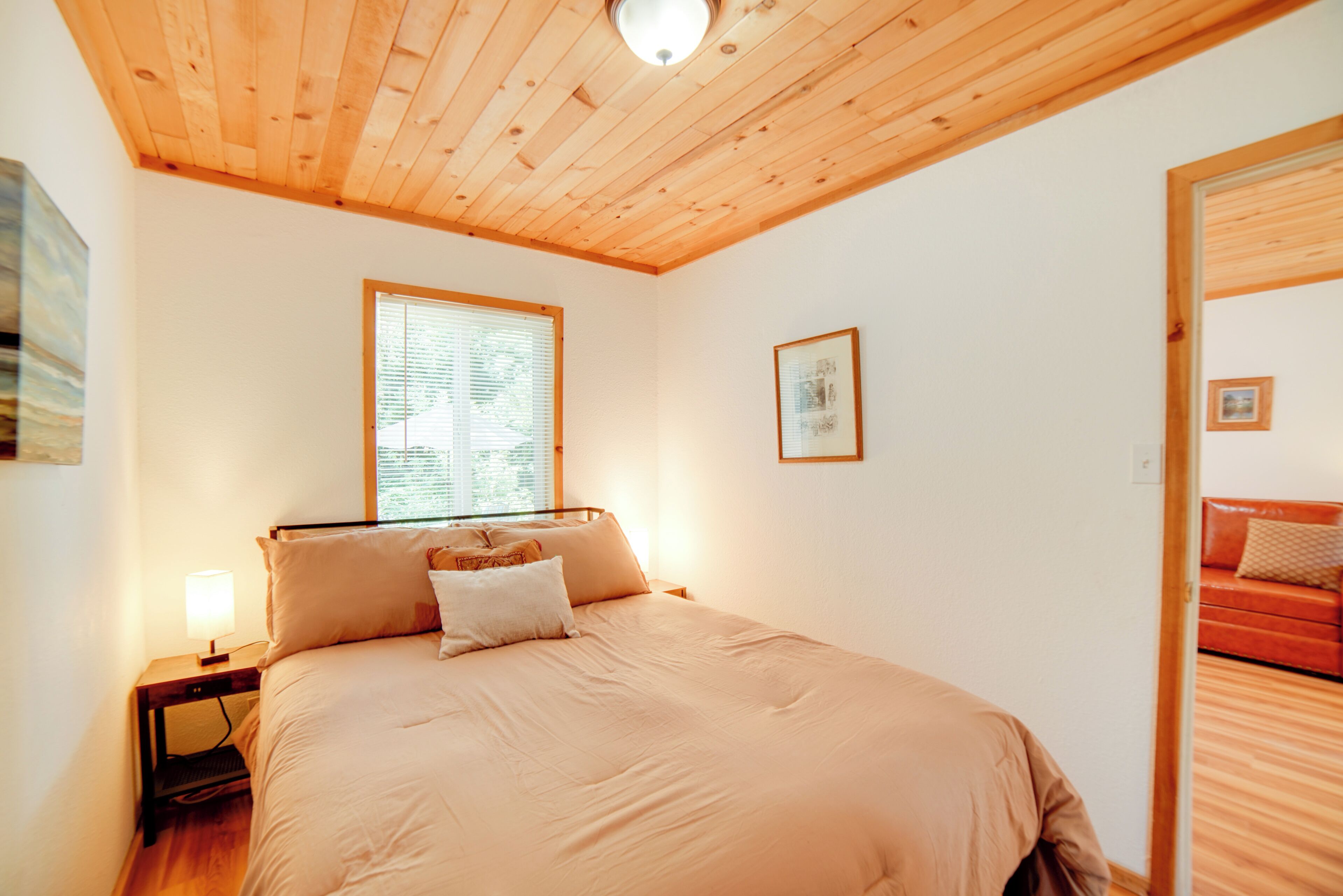 Un dormitorio con techo de madera, una cama con ropa de cama color beige, una mesita de noche con una lámpara, una ventana con persianas y un cuadro en la pared.